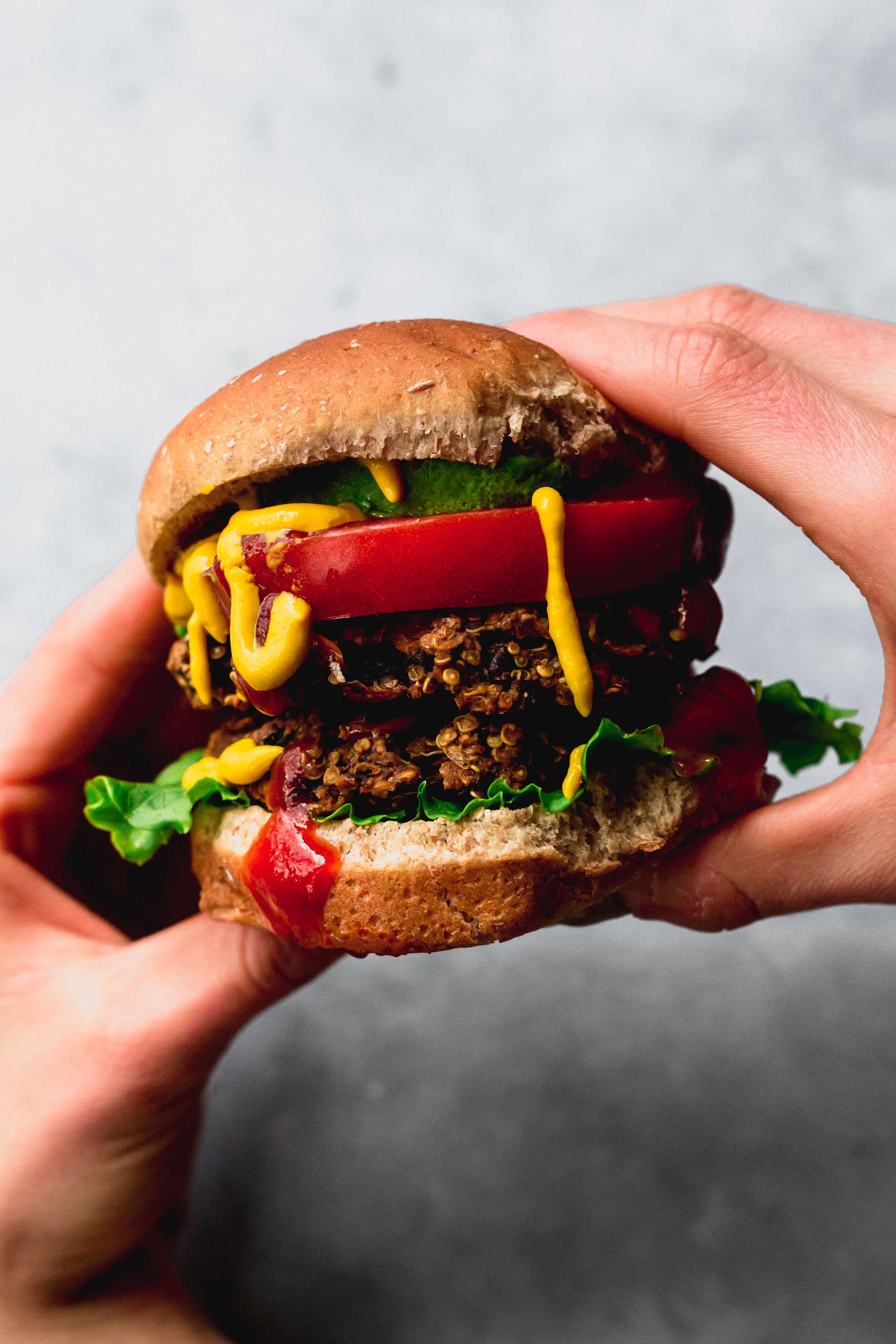 two hands holding a double veggie burger topped with a slice of tomato and a drizzle of mustard