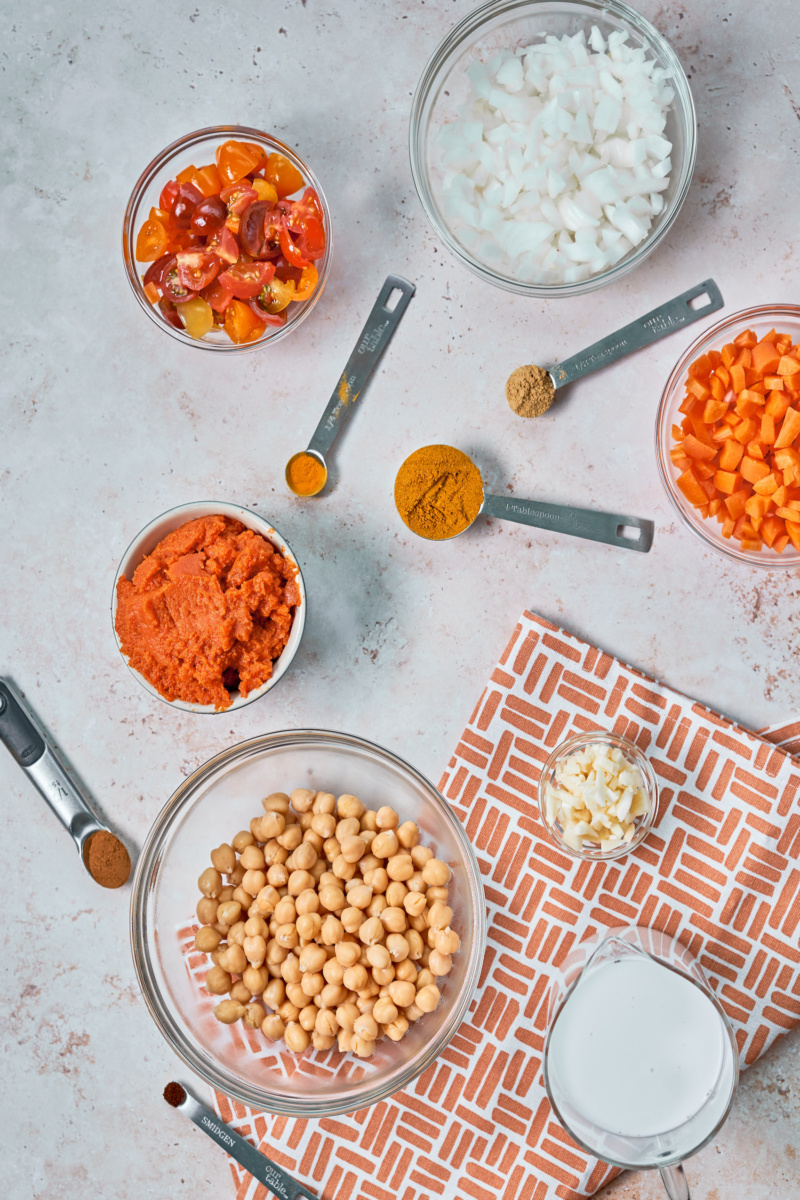 a bowl of onions, a bowl of tomatoes, a bowl of diced carrots, a cup of pureed pumpkin, a bowl of chickpeas, a cup of minced garlic, a measuring cup of coconut and measuring spoons full of spices, all resting on a countertop