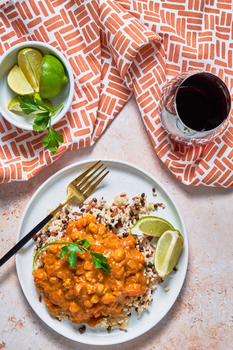 a plate of chickpea pumpkin curry served over a grain blend with lime wedges and a glass of red wine