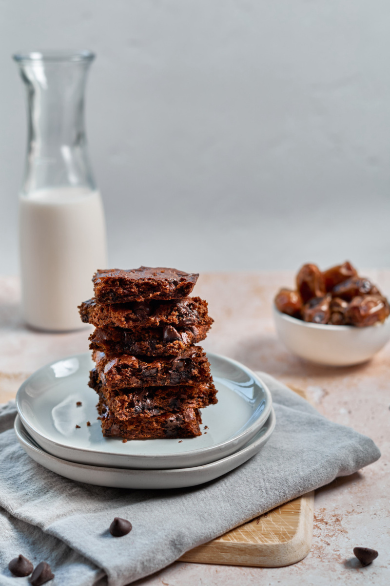 a plate of almond butter chocolate chip blondies stacked on top of one another with a jug of milk and bowl of dates in the background