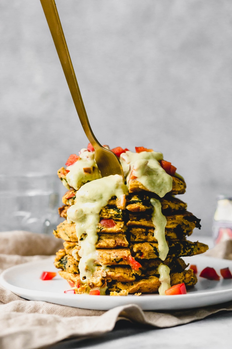 a fork digging into a stack of savory pancakes topped with avocado sauce