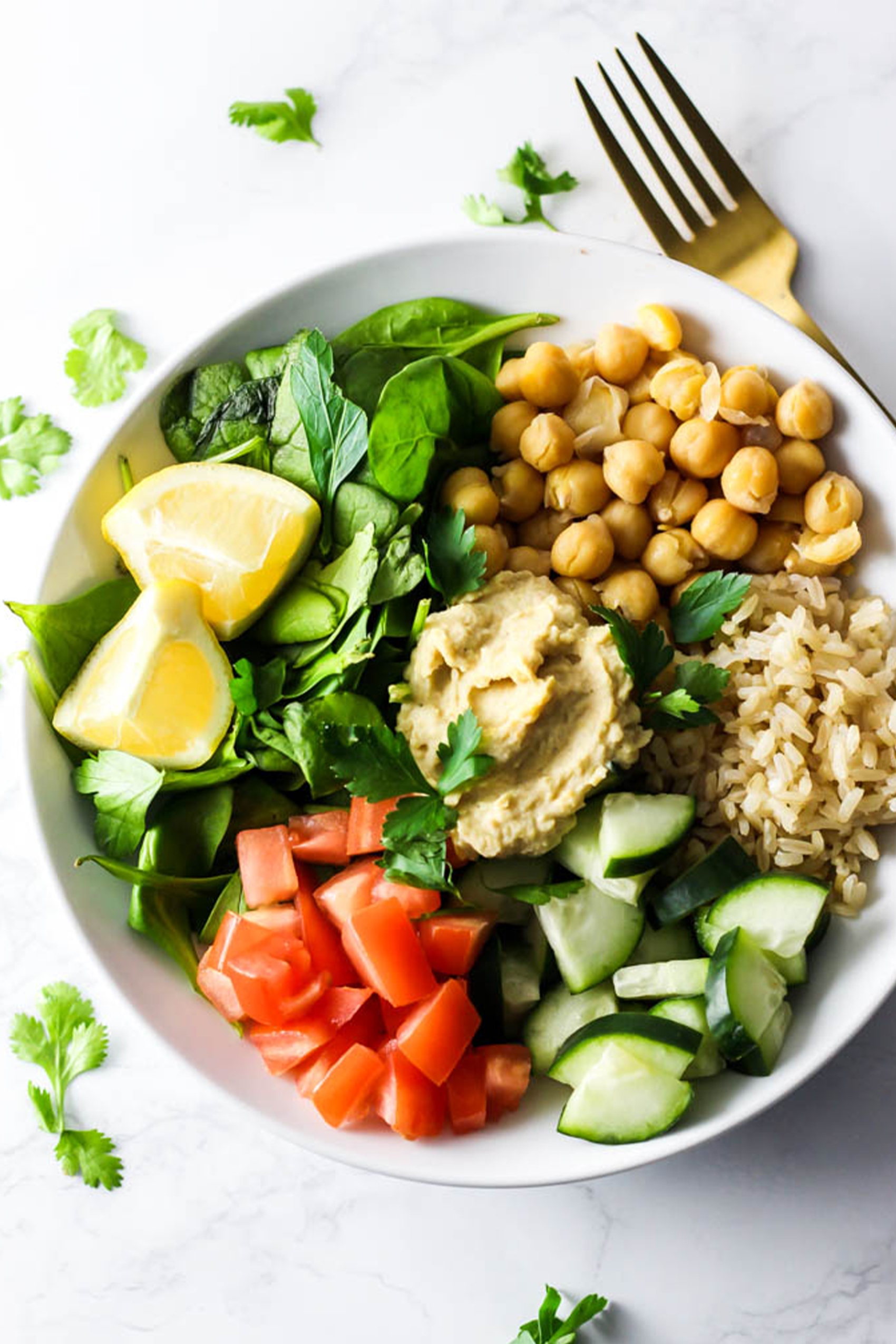 a bowl of spinach served with tomatoes, cucumber, chickpeas, rice, lemon wedges and hummus