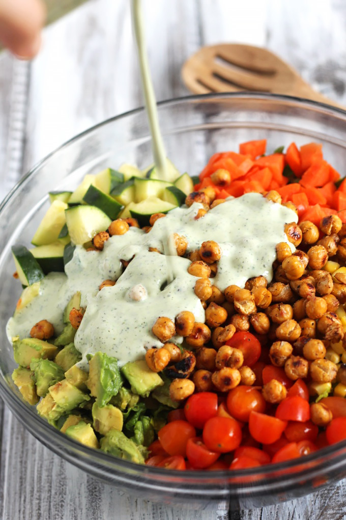 A mixing bowl with avocado, chopped cucumber, cherry tomatoes, carrots and romaine topped with roasted chickpeas and a drizzle of vegan ranch dressing