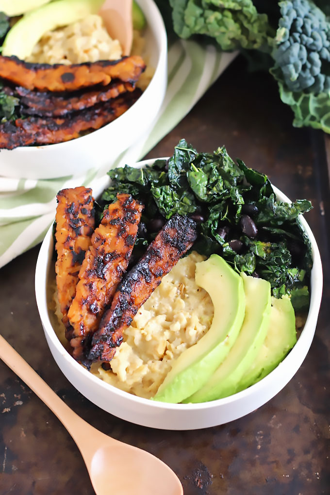 a bowl of oats topped with tempeh bacon, slices of avocado and sauteed kale
