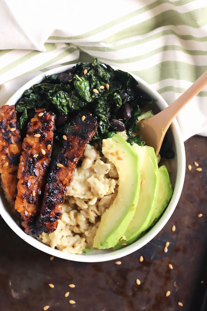 a bowl of vegan oats topped with tempeh bacon strips, avocado slices, and sauteed greens
