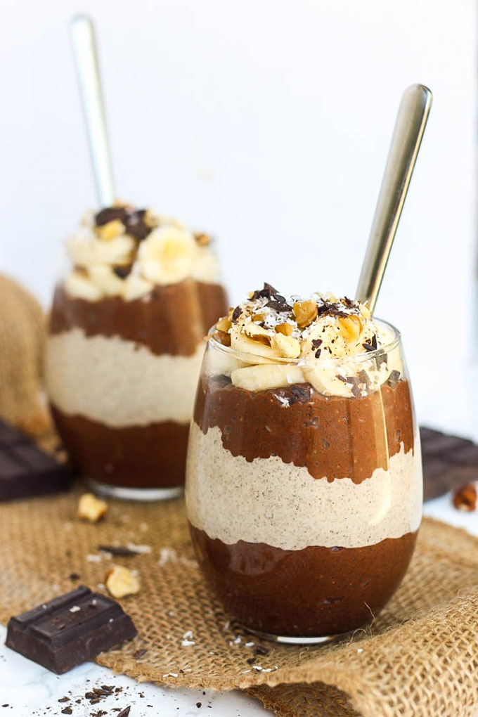 two cups of layered chocolate and peanut butter chia puddings with sliced bananas and chocolate on top