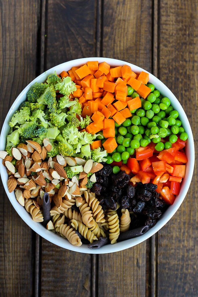 A bowl of vegan pasta salad before it's mixed, containing chopped almonds, raw broccoli, carrots, green peas, red bell pepper, legume pasta and raisins