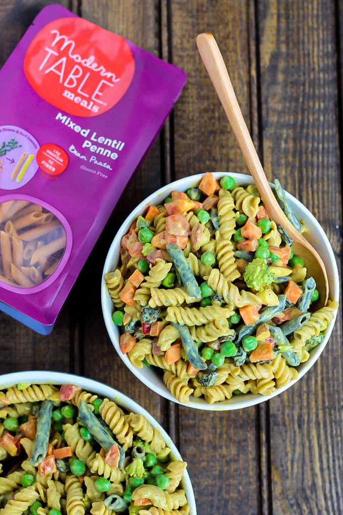 Two bowls of vegan pasta salad with vegetables and a curried sauce served alongside a bag of lentil pasta