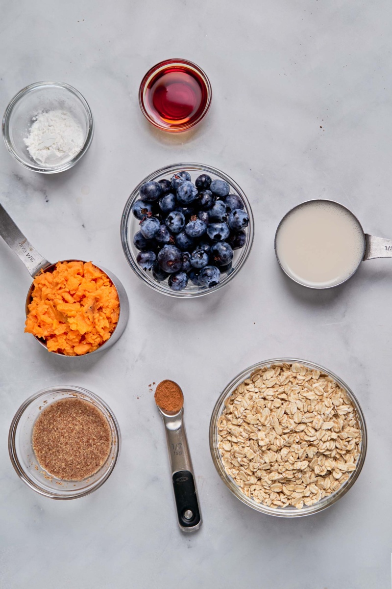 a bowl of oats, a table spoon of cinnamon, a bowl of flax meal, a measuring cup of mashed sweet potato, a bowl of blueberries, a cup of tapioca starch, a cup of maple syrup and a measuring cup of almond milk