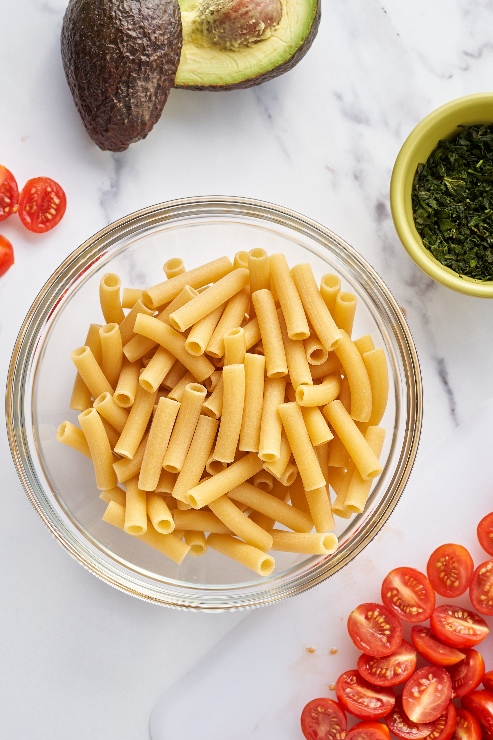 a bowl of dried pasta surrounded by halved cherry tomatoes, a bowl of chopped herbs and an avocado that has been cut in half