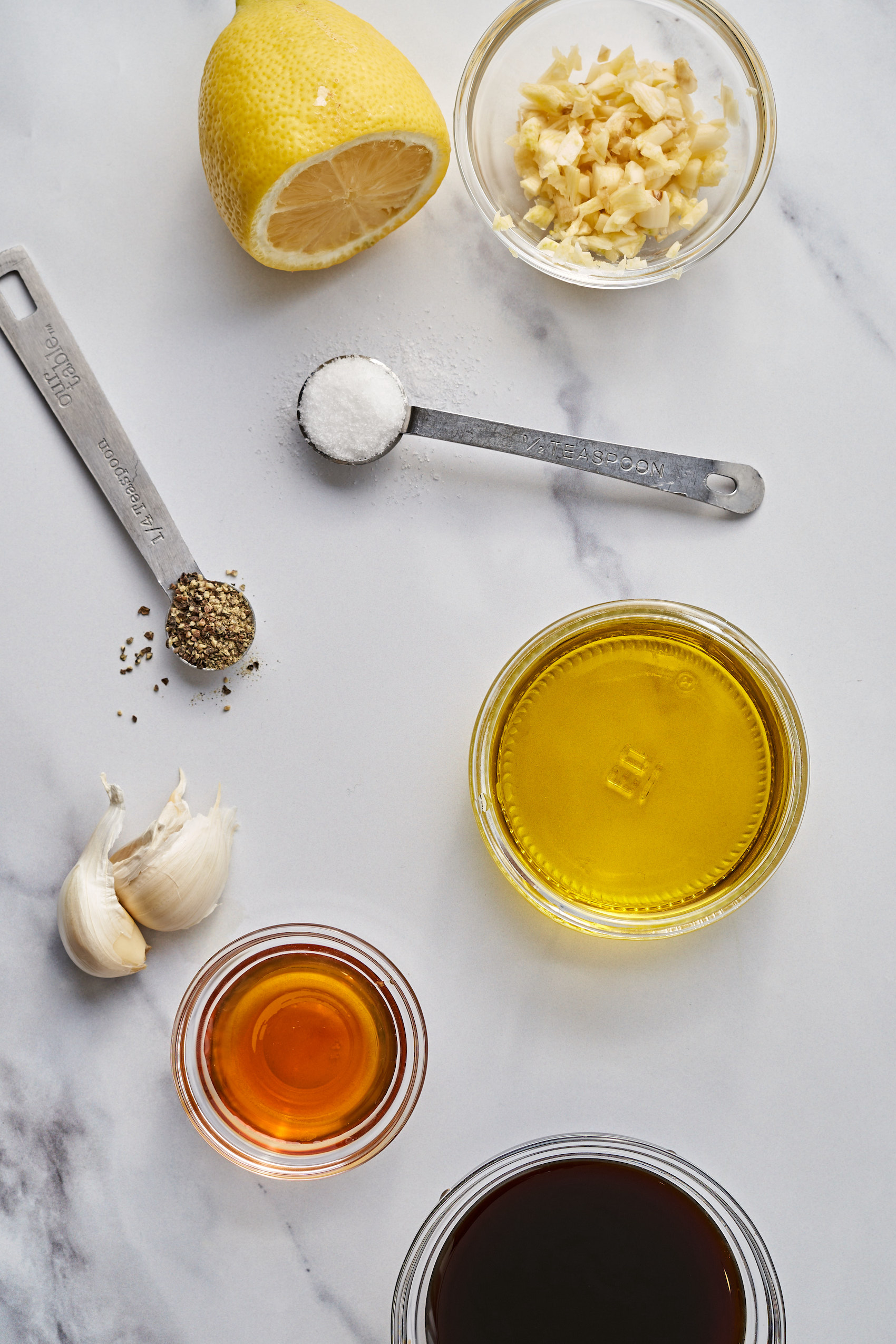 a collection of dressing ingredients, including a small bowl of oil, vinegar, garlic, a half of a lemon and teaspoons of salt and pepper