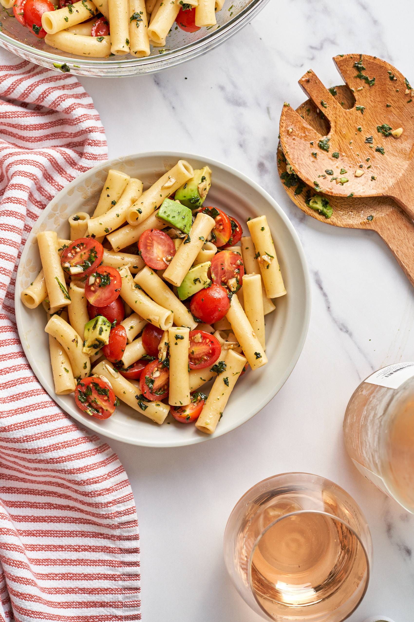 a large bowl of caprese pasta salad next to a wooden salad spoon and a glass of rose wine