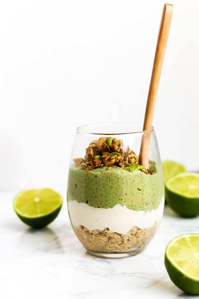 a vegan parfait served in a clear glass featuring lime chia pudding, oat crust and vanilla vegan pudding, all surrounded by halved limes