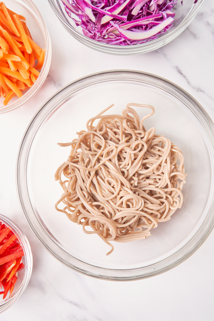 a bowl of cooked soba noodles served next to bowls of fresh sliced vegetables including carrot, bell pepper and purple cabbage