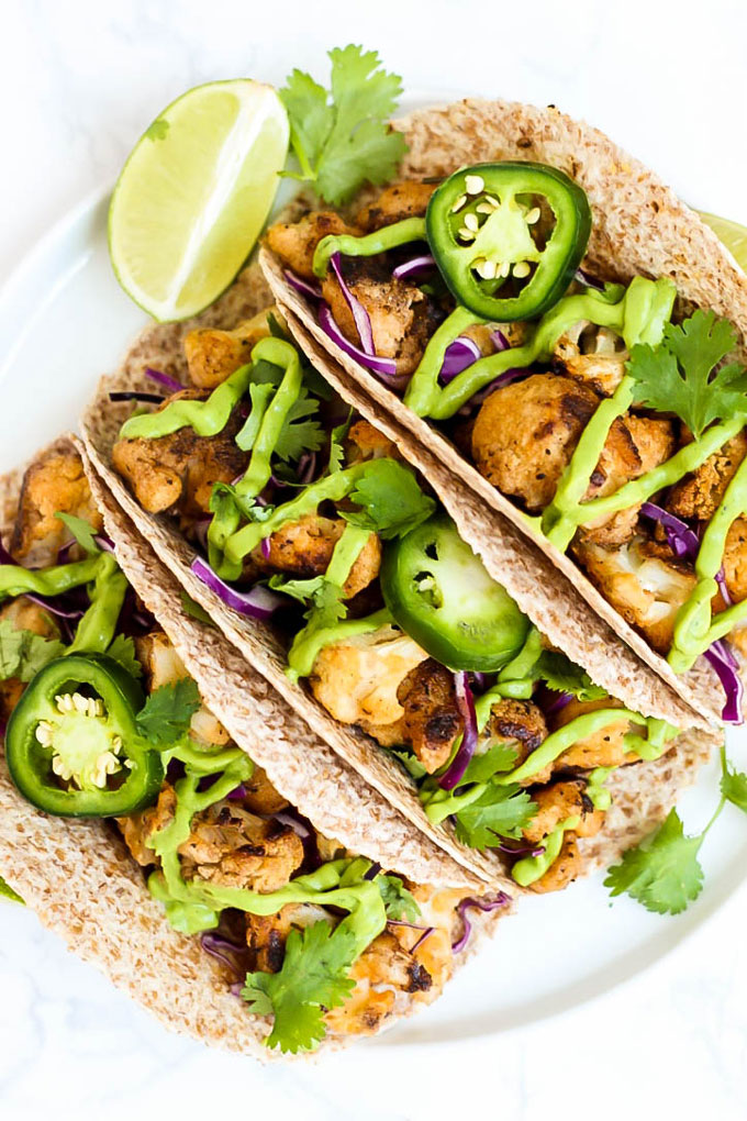 a plate of buffalo cauliflower tacos topped with avocado sauce, sliced jalapenos and lime wedges