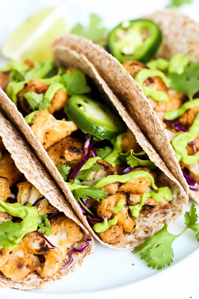 three cauliflower tacos served with fresh lime wedges, sliced jalapenos, and a drizzle of avocado crema