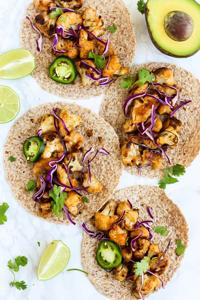 an overhead shot of four tortillas each topped with roasted cauliflower, sliced jalapenos, red cabbage and cilantro