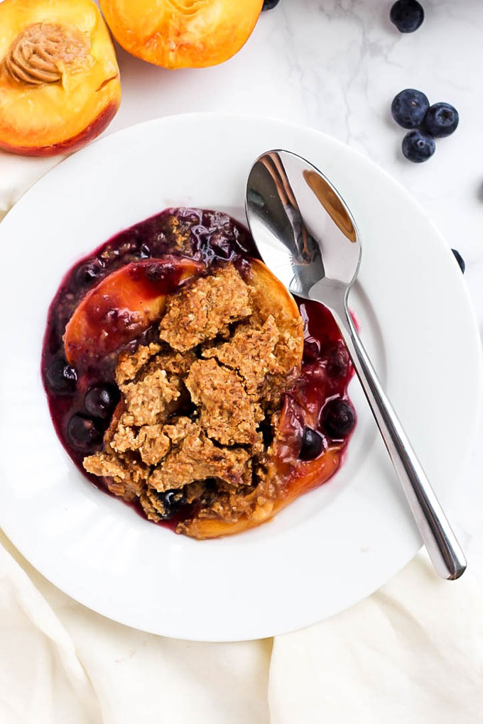 A bowl of blueberry peach cobbler served alongside a halved peach and a few raw blueberries