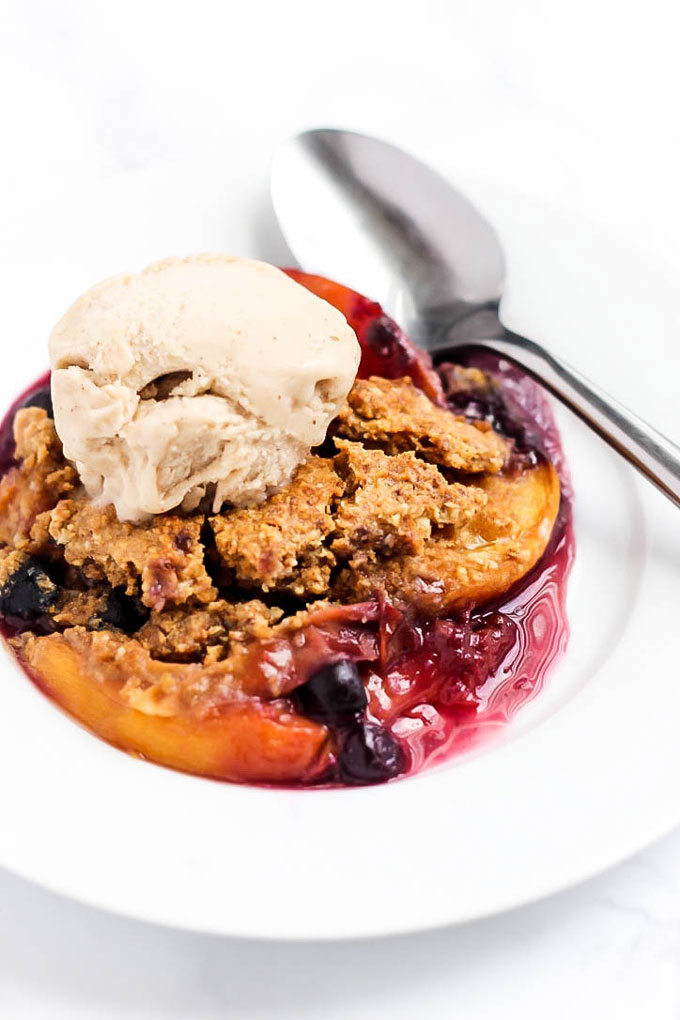 A close up of a white bowl of vegan cobbler made with peaches and blueberries and topped off with a scoop of vegan vanilla ice cream