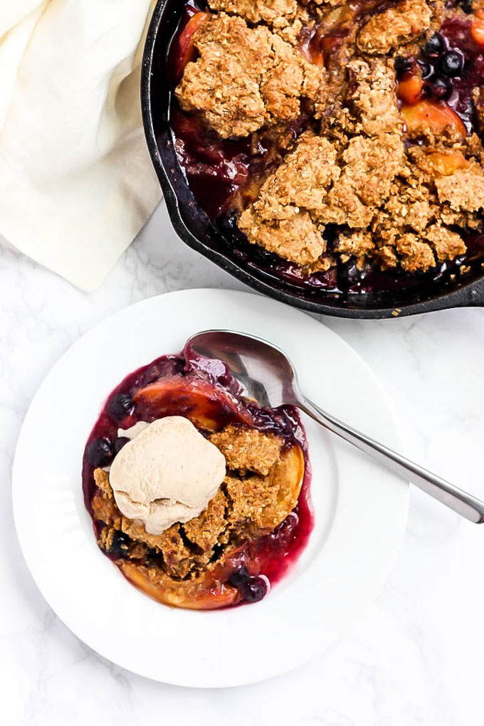 A bowl of blueberry peach cobbler topped with vanilla ice cream alongside the full cast iron skillet of cobbler