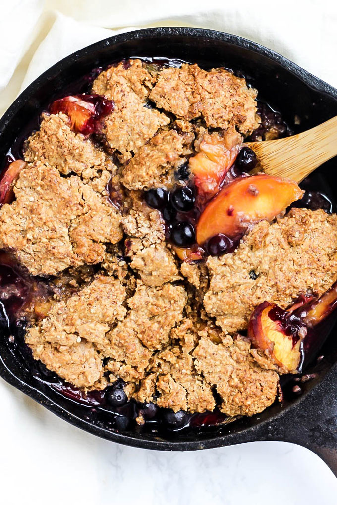A wooden spoon scoops into a cast iron skillet of gluten free blueberry and peach cobbler