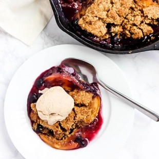 A cast iron skillet of peach and blueberry cobbler next to a serving of the cobbler in a white bowl served with a scoop of vanilla vegan ice cream