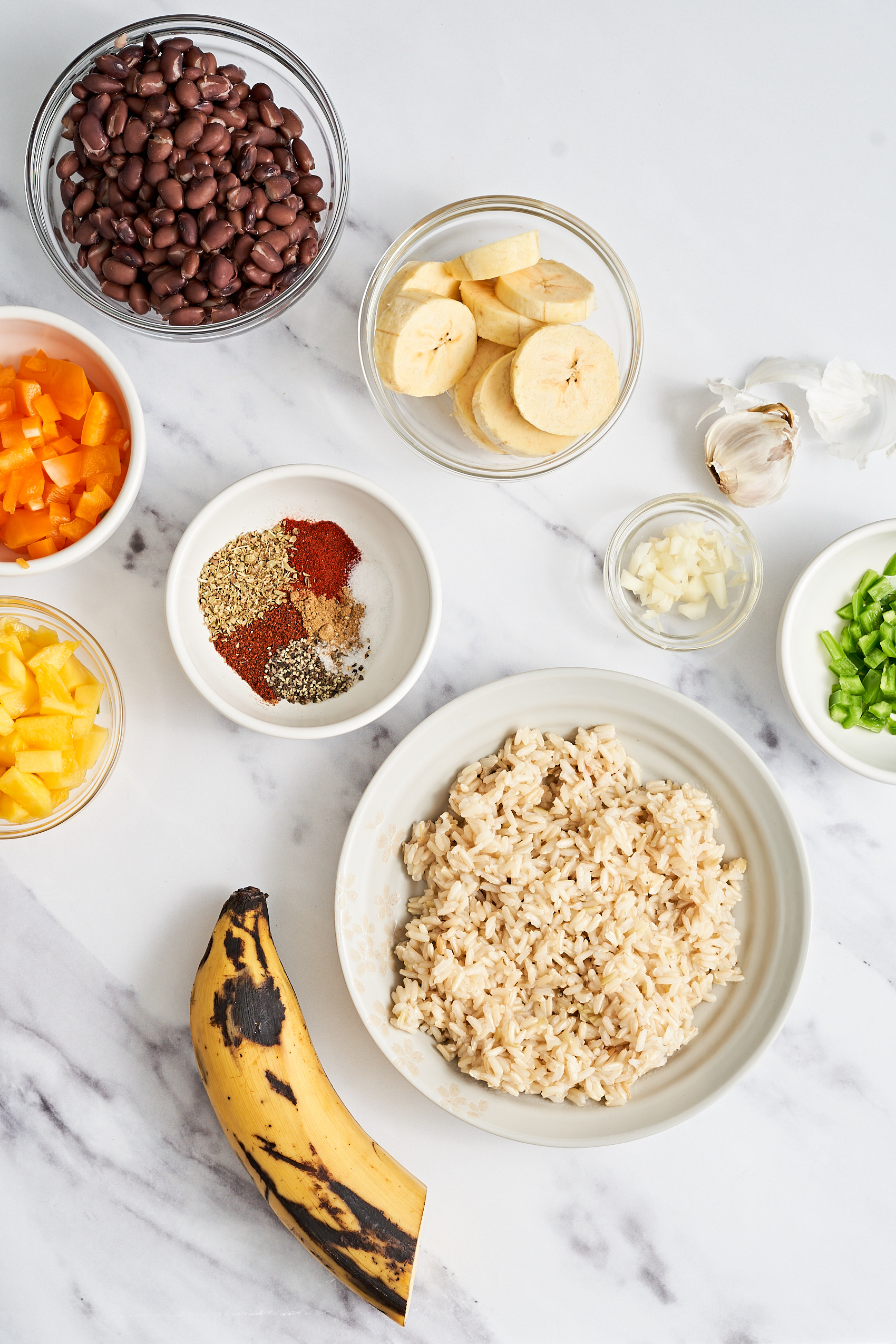 a collection of ingredients arranged on a countertop including a bowl of sliced plantains, a whole plantain, a bowl of rice, chopped green peppers, a bowl of spices, chopped mango and black beans