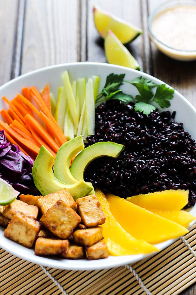 A power bowl filled with black rice, sriracha tofu, mango, vegetables and served with limes