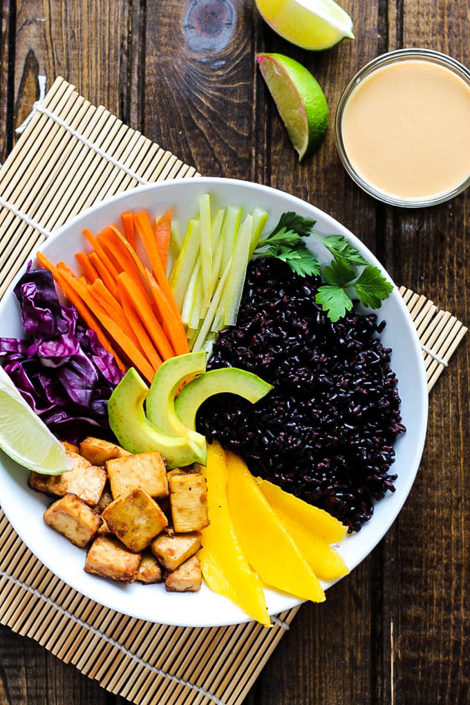 A bowl filled with black rice, mango, tofu, avocado, sliced vegetables and topped with cilantro and a lime wedge