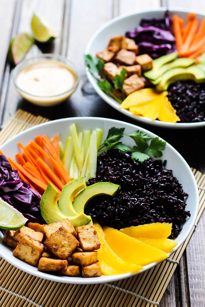 Two power bowls filled with mango, vegetables, avocado, tofu and black rice served with lime wedges and tahini sriracha sauce