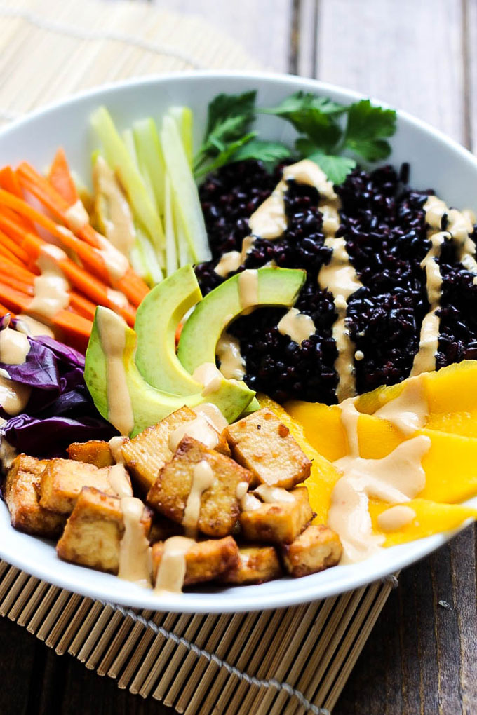 A bowl filled with sliced vegetables, black rice, tofu, avocado and a tahini sriracha sauce