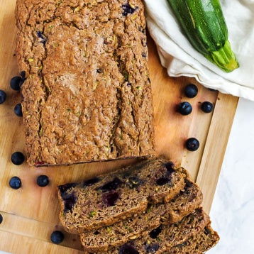 An overhead shot of a blueberry zucchini loaf and four cut slices resting on a wooden cutting board, surrounded by blueberries and a zucchini