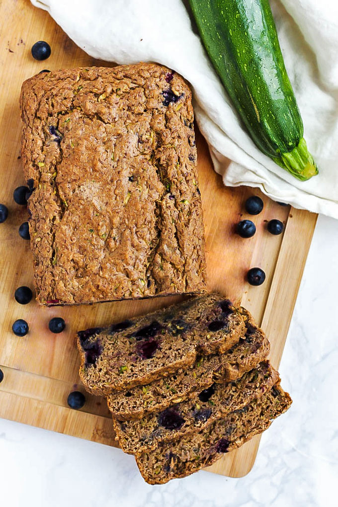 An overhead shot of a blueberry zucchini loaf and four cut slices resting on a wooden cutting board, surrounded by blueberries and a zucchini