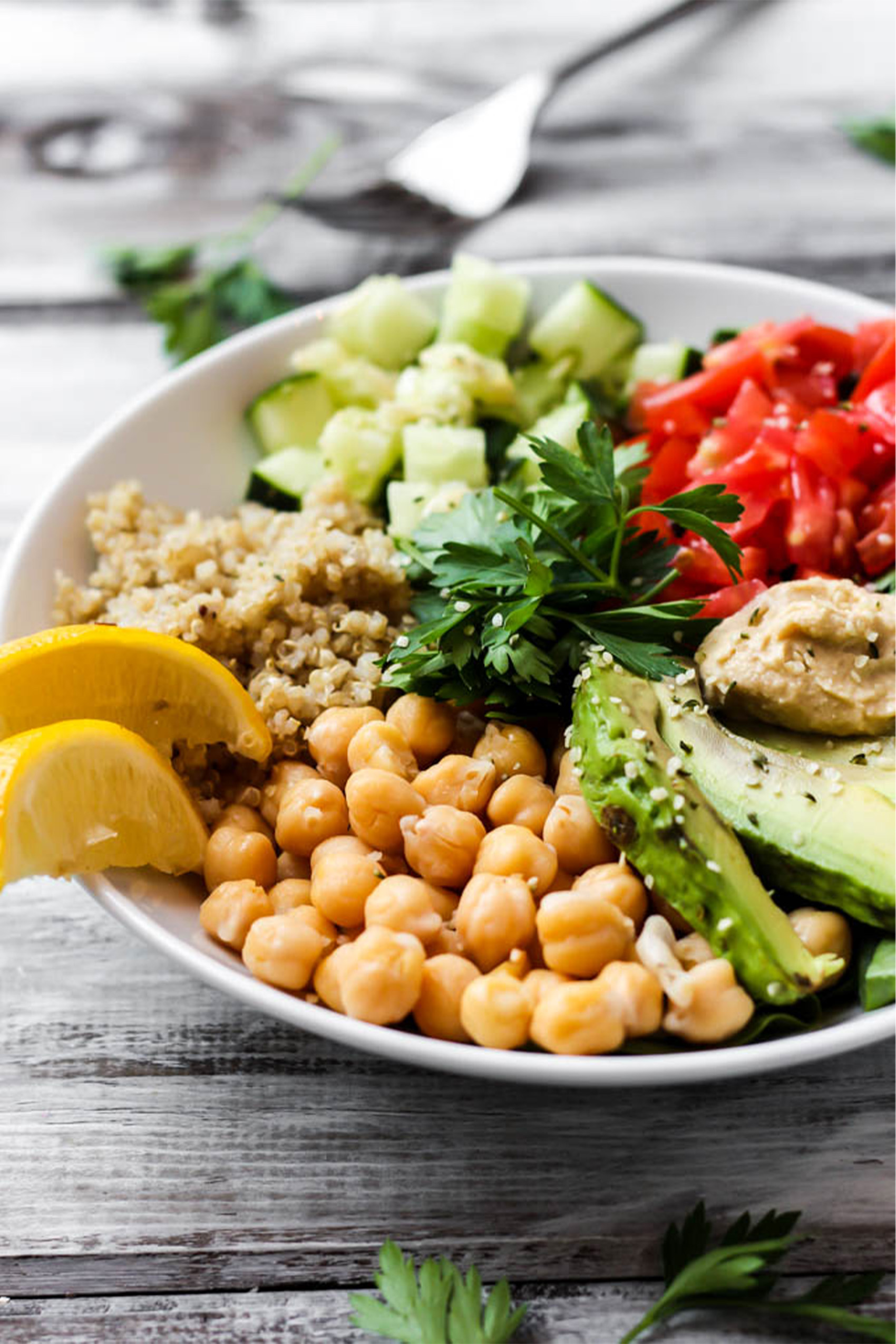 a greek quinoa bowl with chickpeas, sliced avocado, tomatoes, cucumber, quinoa, parsley, hummus and lemon wedges