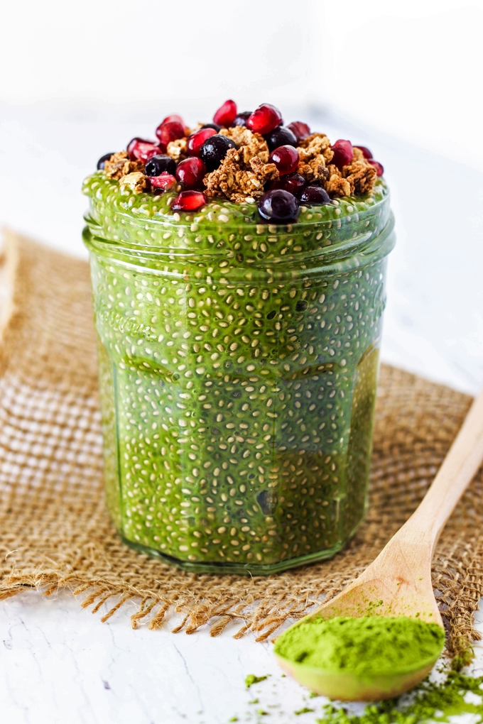 a jar filled with a matcha chia pudding topped with blueberries, pomegranate and granola