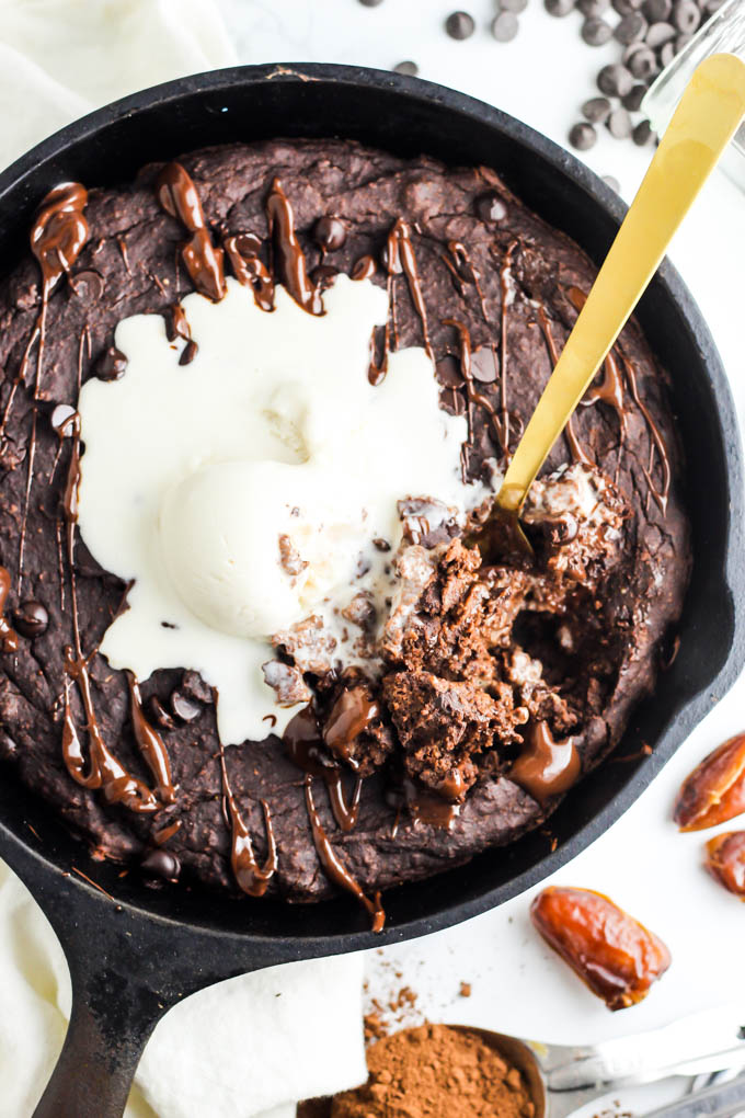 An overhead shot of a gold fork scooping into a brownie baked in a cast iron skillet. The brownie is topped with melted chocolate and a scoop of vanilla vegan ice cream