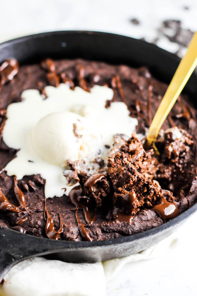 A gold fork digging into a vegan brownie served in a cast iron skillet with a scoop of vanilla dairy-free ice cream.
