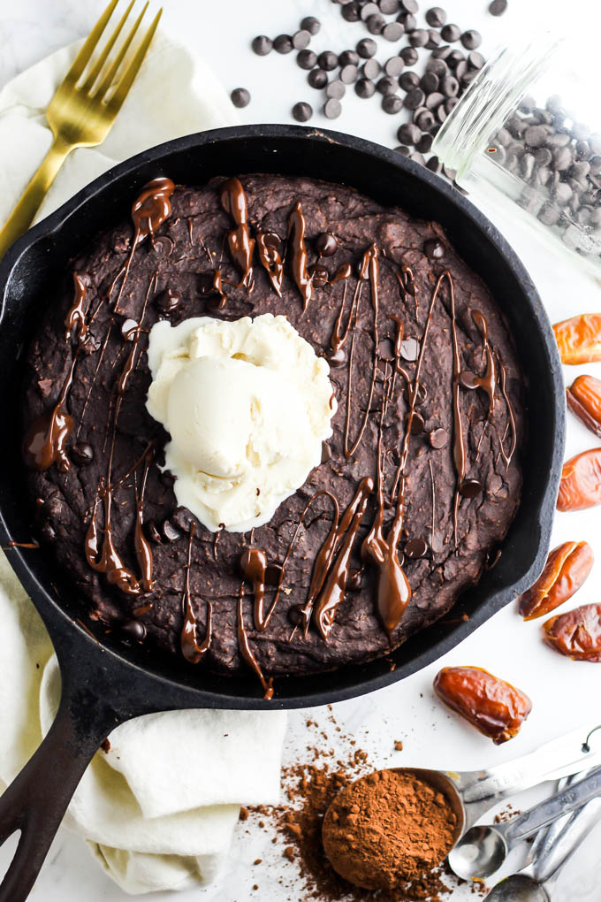 A bird's-eye shot of a cast iron skillet brownie. The skillet is surrounded by a gold fork, a jar of chocolate chips spilling onto the counter, dates and a tablespoon of cocoa powder. The brownie is topped with vanilla vegan ice cream and a drizzle of chocolate