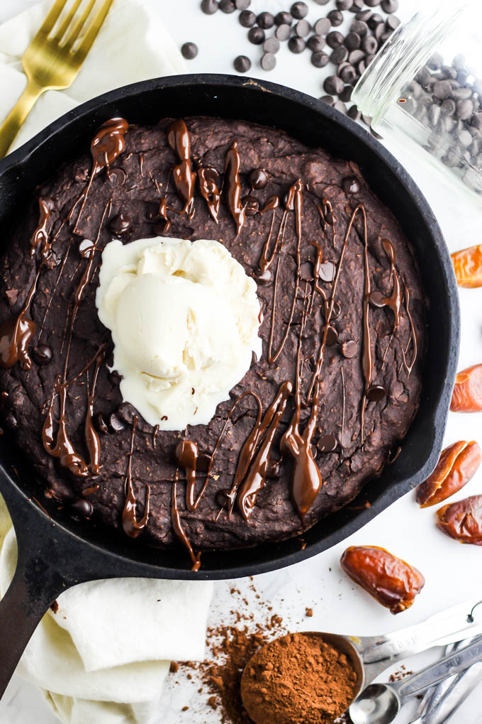 A brownie baked in a cast iron skillet is served with a scoop of vanilla dairy-free ice cream and a drizzle of melted chocolate. The pan is served on a white countertop, surrounded by a tablespoon of cocoa powder, dates, a jar of chocolate chips and a gold fork