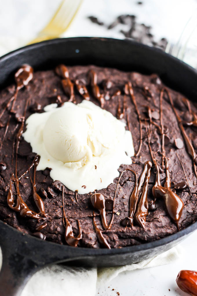 A vegan brownie baked in a cast iron skillet drizzled with melted chocolate and topped off with a scoop of vanilla vegan ice cream