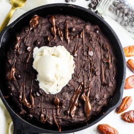 A close up of a baked brownie served in a cast iron skillet, drizzled with melted chocolate and topped with vanilla dairy-free ice cream