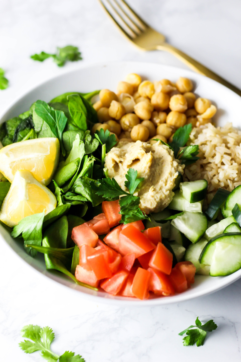 a bowl filled with spinach, brown rice, cucumber, tomatoes, and chickpeas topped off with hummus, parsley and a lemon wedge