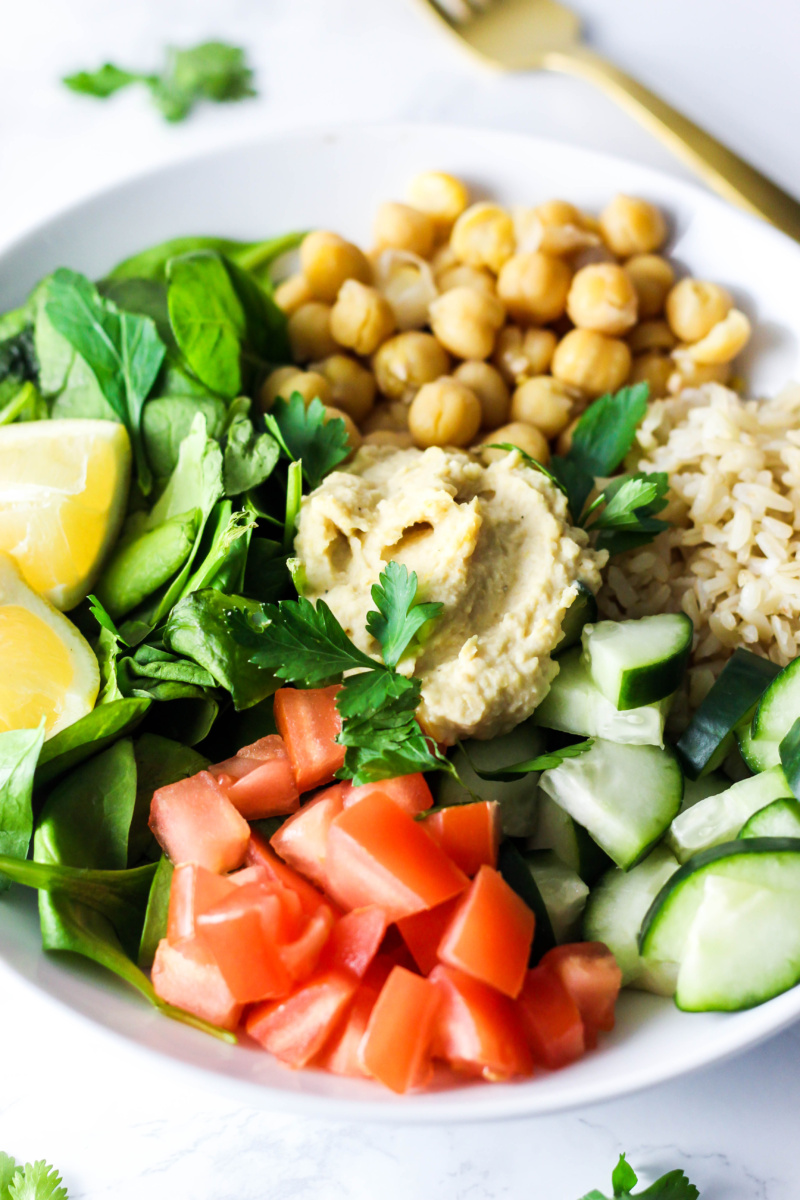 a salad filled with chickpeas, brown rice, chopped tomatoes, cucumber, spinach, parsley, a dollop of hummus and two lemon wedges