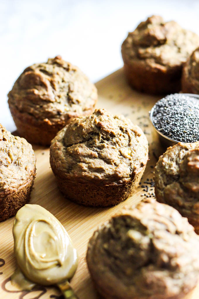 a batch of banana muffins on a cutting board served with a spoonful of almond butter and a measuring cup of chia seeds