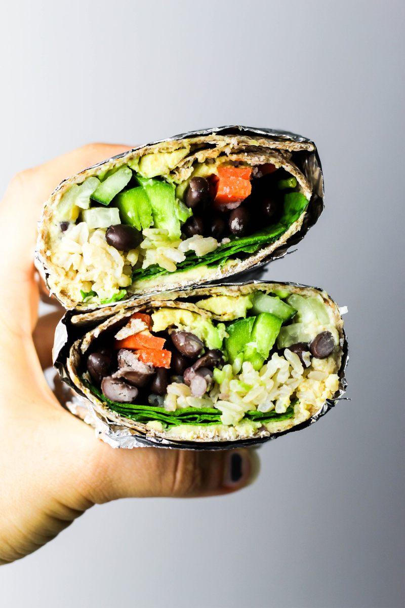 a hand holds two halves of a vegan wrap filled with greens, rice, veggies and black beans