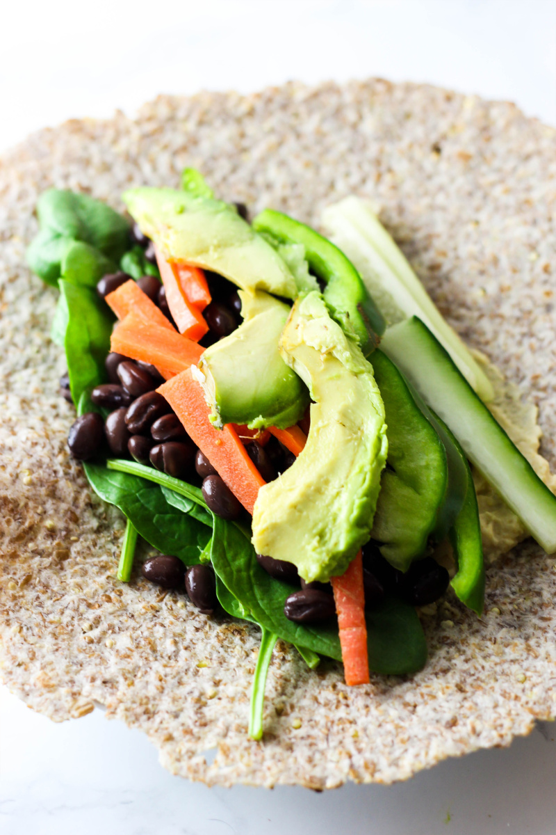 avocado, sliced carrots, bell pepper, greens and black beans lay on top of a tortilla