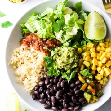 a lunch bowl filled with black beans, brown rice, salsa, lettuce, a wedge of lime, corn, peppers and a dollop of guacamole
