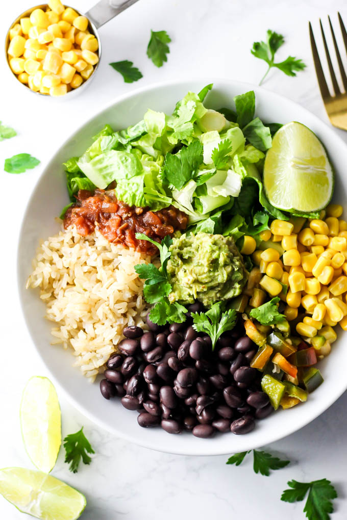 a lunch bowl filled with black beans, brown rice, salsa, lettuce, a wedge of lime, corn, peppers and a dollop of guacamole