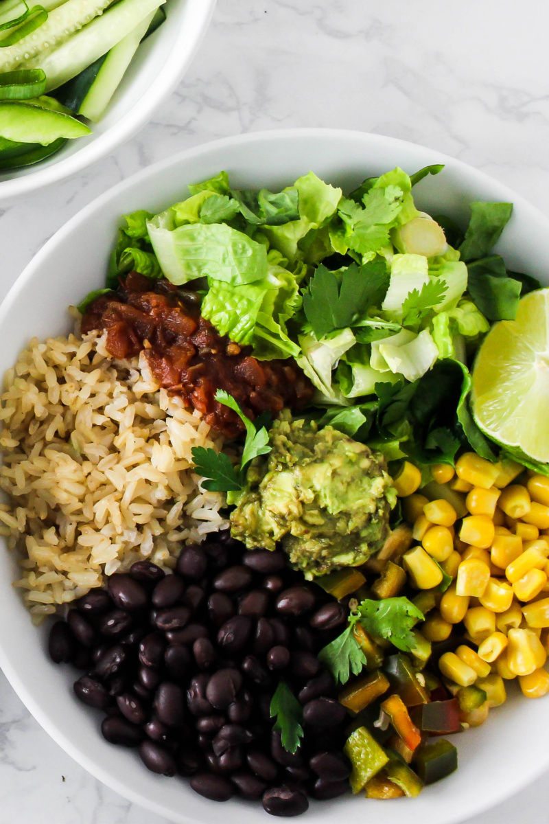 a burrito bowl with black beans, brown rice, salsa, lettuce, corn, peppers, cilantro and guacamole