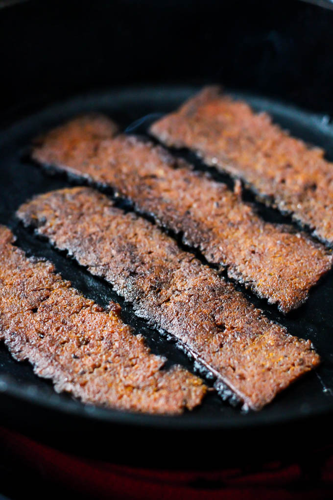 Four slices of vegan bacon being fried in a cast iron skillet